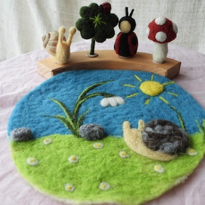 Könnte beinhalten: Eine kreisförmige, nadelgefilzte Matte mit blauem Himmel, gelber Sonne, grünem Gras, weißen Blumen und einer Schnecke. Auf einem geschwungenen Holzständer stehen eine gefilzte Schnecke, ein grüner Klee mit Marienkäfer, eine Marienkäferfigur und ein Pilz.