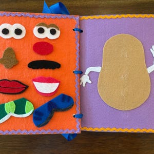 Può includere: Un libro di attività in feltro con una pagina arancione che presenta vari tratti del viso, tra cui occhi, naso, baffi, labbra e occhiali. La pagina viola adiacente mostra una figura color marrone chiaro a forma di patata con braccia bianche.