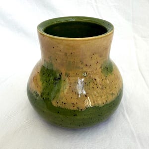 May include: A ceramic vase with a wide, rounded body and a flared rim. The surface features a two-tone glaze with a tan upper section and a deep green lower section, both decorated with small, dark brown speckles throughout the entire exterior.