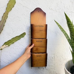 May include: A vertical wooden wall organiser with three stacked compartments. Each compartment is labelled with black text reading LETTERS, BILLS, and MISC. The wood features a light brown centre with darker, burnt-style edges and three small metal hooks at the bottom.