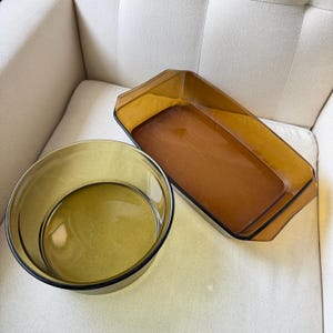 Puede incluir: Un cuenco redondo de cristal verde oliva y una fuente de horno rectangular de cristal ámbar. Ambas piezas están hechas de cristal transparente y liso con diseños sencillos y funcionales, con aberturas superiores y bordes moldeados.