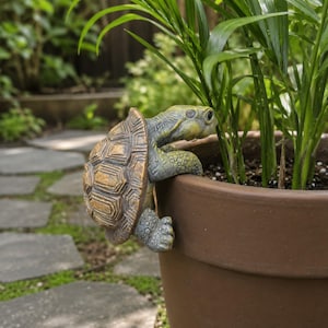 Könnte beinhalten: Eine dekorative Schildkrötenfigur in Braun-, Grün- und Gelbtönen, die sich am Rand eines braunen Blumentopfs festhält. Die Schildkröte hat einen detaillierten Panzer und befindet sich in der Nähe von grünen Pflanzen, was auf einen Garten hindeutet.