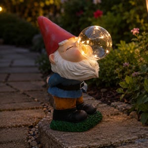 Può includere: Una statua di gnomo da giardino stravagante con un cappello rosso, una giacca blu e una barba bianca. Lo gnomo sembra soffiare una bolla, che in realtà è un globo trasparente pieno di luci calde e scintillanti. La statua è posta su un sentiero di pietra.