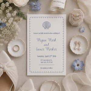 May include: A white wedding invitation card with a blue shell icon, featuring the text Megan Mark and Tanner Marker, Saturday, April 22nd, 2030, 123 Anywhere St., Any City, ST 12345, and Reception to Follow, surrounded by a perfume bottle, rings, and jewelry.