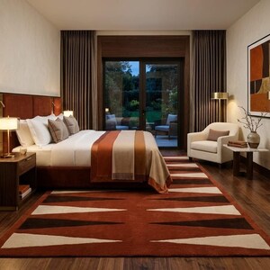 Op de afbeelding: Een slaapkamer met een groot bed met een roestkleurig gestoffeerd hoofdeinde, wit beddengoed en een bruin- en crèmekleurige sprei. Een vloerkleed met geometrisch patroon in roest, bruin en crème ligt op de donkere houten vloer.
