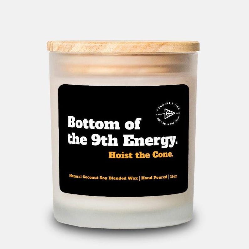 May include: A cylindrical candle in a frosted glass jar with a wooden lid. The black label features the text "Bottom of the 9th Energy. Hoist the Cone." and "Natural Coconut Soy Blended Wax | Hand Poured | 11oz."
