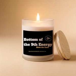 May include: A lit white candle in a frosted glass jar with a wooden lid. The candle has a black label with the text "Bottom of the 9th Energy" and "Hoist the Cone."
