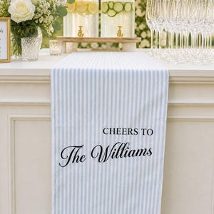Puede incluir: Un camino de mesa con rayas verticales en azul claro y blanco que presenta el texto impreso en negro CHEERS TO The Williams. El camino de tela cuelga sobre el borde de una mesa blanca, mostrando un diseño rectangular de líneas rectas.