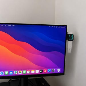 Może przedstawiać: Monitor komputera z kolorowym, abstrakcyjnym tłem. Biały smartwatch, wyświetlający godzinę na zielono, jest przymocowany do ściany obok monitora.