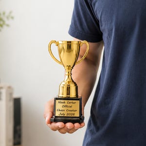 May include: A gold-colored trophy with two handles mounted on a black square base. The base features a plaque with the engraved text: Noah Carter, Official, Chaos Creator, July 2026. The trophy has a polished, reflective metallic finish.