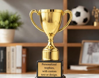 May include: A shiny gold-coloured trophy cup with two handles, mounted on a black square base. The base features a gold-toned plaque with the black text: Personalized trophies, with custom text or design. The trophy has a smooth, reflective finish.