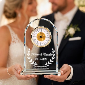 May include: An arched clear glass clock featuring a gold-rimmed circular face with Roman numerals. The glass is etched with the names Philippe & Camille, the date 08.10.2026, a laurel wreath design, and the text You are my yesterday, today, and every tomorrow.