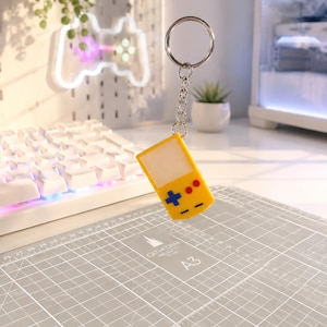 May include: A yellow handheld gaming console keychain featuring a white screen area, a blue directional pad, and two red circular buttons. The keychain is attached to a silver metal split ring by a short silver chain, suspended in the air.