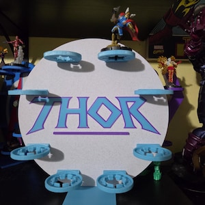 以下が含まれることがあります： 大きな青と紫の文字で「THOR」と書かれた円形の白いディスプレイスタンド。スタンドの前面には小さなアクションフィギュアを保持するための6つの水色の円形プラットフォームがあり、そのうち1つにフィギュアが配置されています。