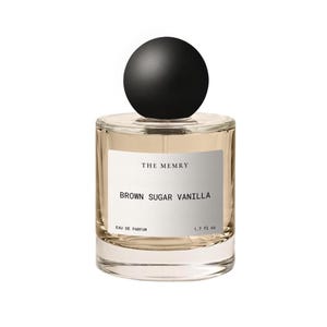 May include: A clear glass perfume bottle with a round black cap. The white label features the text THE MEMRY, BROWN SUGAR VANILLA, EAU DE PARFUM, and 1.7 fl oz. The bottle contains a light amber liquid and has a cylindrical shape.