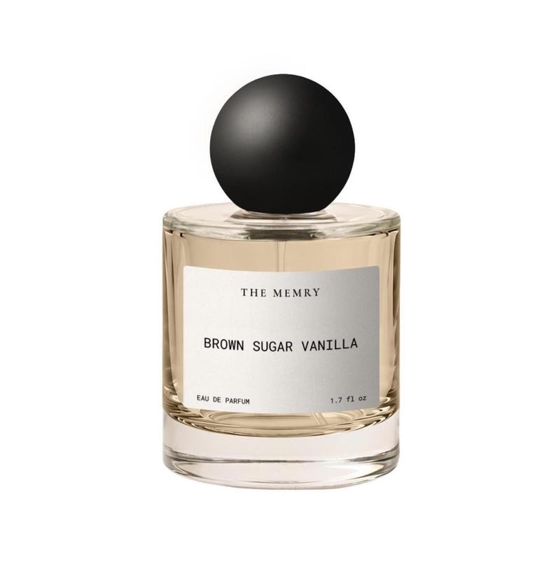May include: A clear glass perfume bottle with a round black cap. The white label features the text THE MEMRY, BROWN SUGAR VANILLA, EAU DE PARFUM, and 1.7 fl oz. The bottle contains a light amber liquid and has a cylindrical shape.