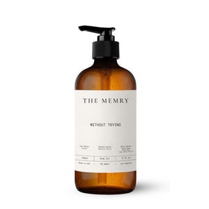 May include: An amber-colored glass bottle with a black pump dispenser containing body oil. The white label features the text THE MEMRY, WITHOUT TRYING, and lists scent notes, 236ml, 8 fl oz, Made in USA, and Los Angeles in black lettering.