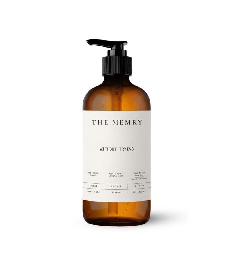 May include: An amber-colored glass bottle with a black pump dispenser containing body oil. The white label features the text THE MEMRY, WITHOUT TRYING, and lists scent notes, 236ml, 8 fl oz, Made in USA, and Los Angeles in black lettering.