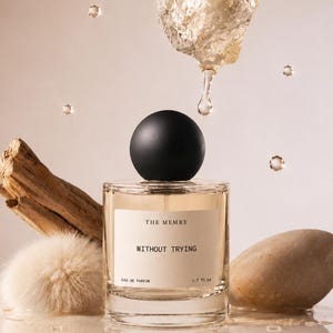 May include: A clear glass perfume bottle with a round black cap, containing light amber liquid. The label reads THE MEMRY WITHOUT TRYING EAU DE PARFUM 1.7 fl oz. It is surrounded by a piece of driftwood, a fluffy beige pom-pom, and a smooth stone.