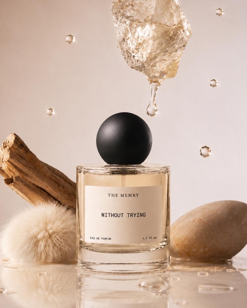 May include: A clear glass perfume bottle with a round black cap, containing light amber liquid. The label reads THE MEMRY WITHOUT TRYING EAU DE PARFUM 1.7 fl oz. It is surrounded by a piece of driftwood, a fluffy beige pom-pom, and a smooth stone.