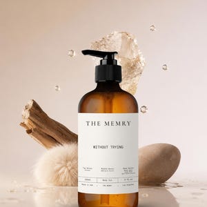 May include: An amber glass bottle with a black pump dispenser containing body oil. The white label features the text THE MEMRY, WITHOUT TRYING, and lists notes, 236ml, and 8 fl oz. The bottle is surrounded by a piece of driftwood, a stone, and a fluffy sphere.