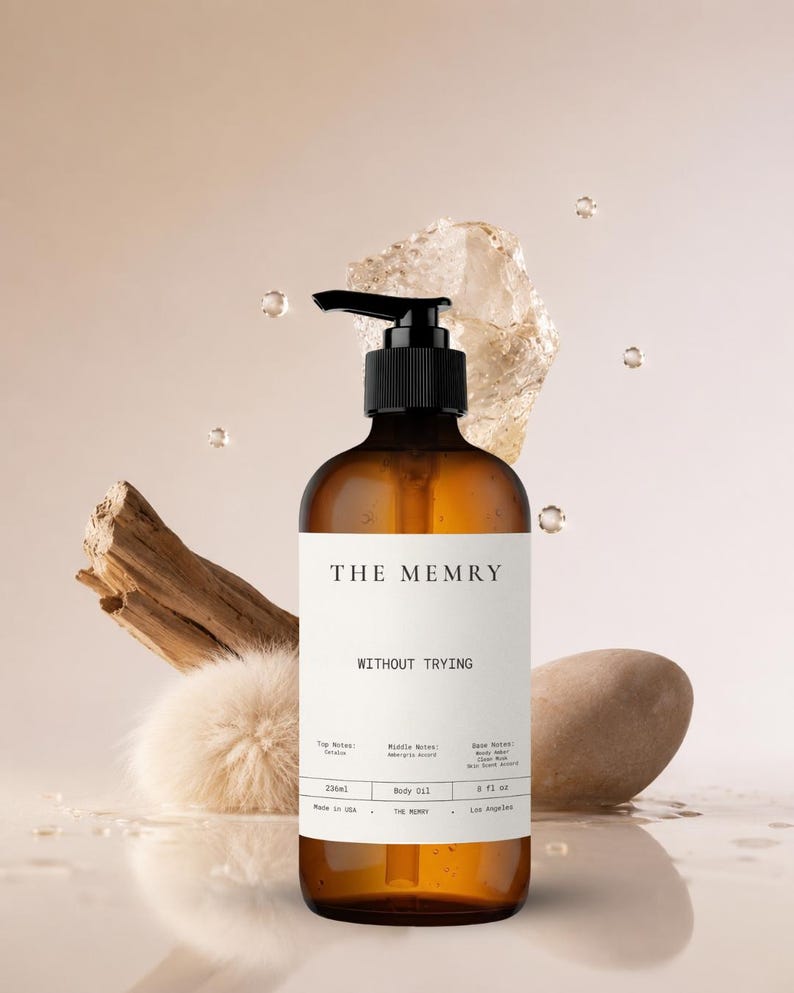 May include: An amber glass bottle with a black pump dispenser containing body oil. The white label features the text THE MEMRY, WITHOUT TRYING, and lists notes, 236ml, and 8 fl oz. The bottle is surrounded by a piece of driftwood, a stone, and a fluffy sphere.