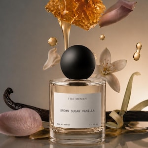 May include: A clear glass perfume bottle with a black spherical cap and a white label reading THE MEMRY BROWN SUGAR VANILLA EAU DE PARFUM 1.7 fl oz. The bottle is surrounded by floating honeycomb, golden liquid, vanilla beans, and white flower petals.