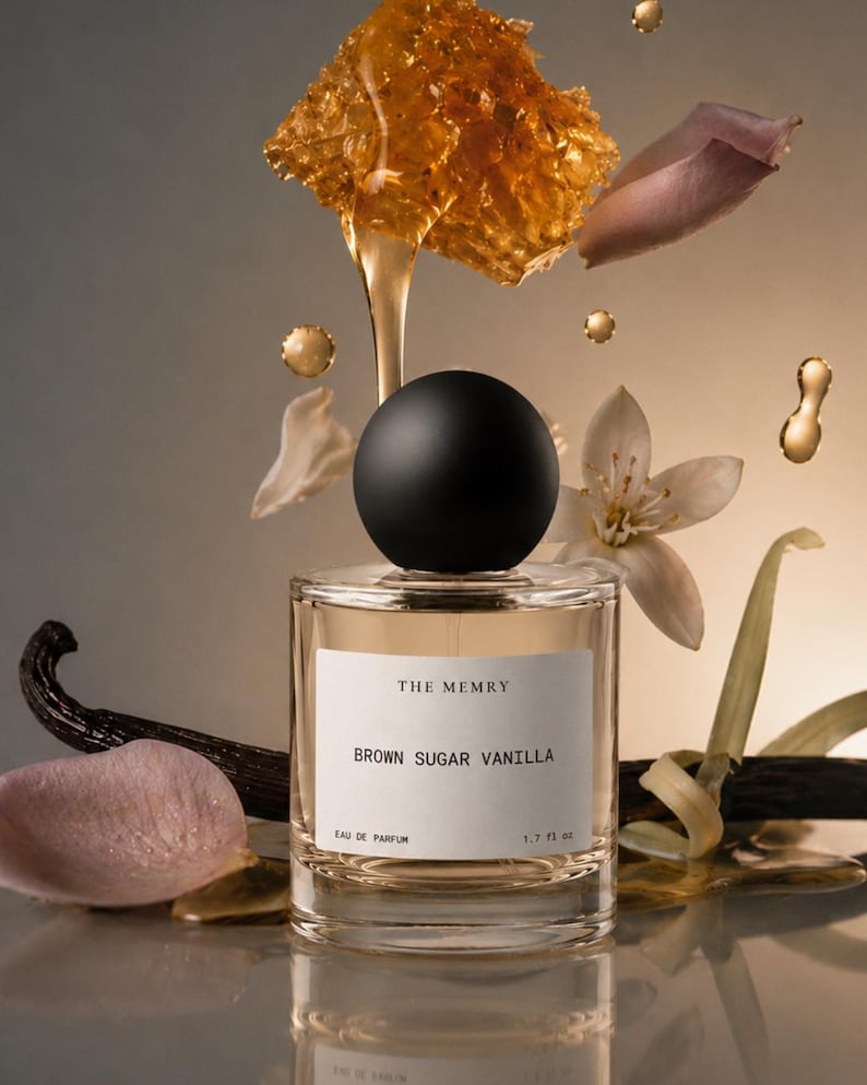 May include: A clear glass perfume bottle with a black spherical cap and a white label reading THE MEMRY BROWN SUGAR VANILLA EAU DE PARFUM 1.7 fl oz. The bottle is surrounded by floating honeycomb, golden liquid, vanilla beans, and white flower petals.