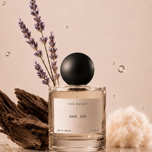 May include: A clear glass perfume bottle with a black spherical cap, featuring a white label that reads THE MEMRY RARE AIR EAU DE PARFUM 1.7 fl oz. The bottle is surrounded by sprigs of purple lavender, a piece of dark wood, and white fiber.