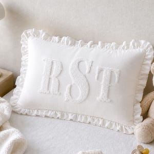 May include: A rectangular white throw pillow featuring a ruffled border and the letters RST embroidered in a raised, textured white font across the center. The pillow is made of a solid white fabric and has a soft, decorative appearance.