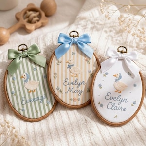 May include: Three oval wooden embroidery hoops featuring embroidered ducklings, floral accents, and names. The left hoop has green stripes and a green bow, the centre has yellow stripes and a blue bow, and the right is white with a white bow.