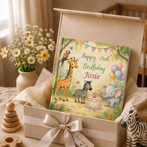 May include: A square gift box containing a personalized book with a jungle theme. The cover features a giraffe, zebra, elephant, and parrot with the text Happy 2nd Birthday Josie. The box is tied with a satin ribbon and includes a gift tag.