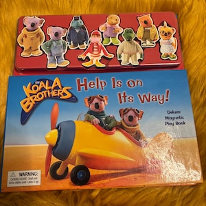 May include: The Koala Brothers Help Is on Its Way! deluxe magnetic play book featuring a yellow aeroplane with two koala characters in flight gear. Above the book is a red metal tin containing eight die-cut magnetic character figures.
