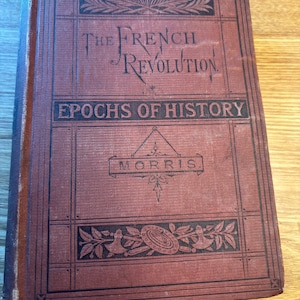 May include: A vintage book titled "The French Revolution" with "Epochs of History" printed below. The cover is a deep red with black decorative elements and text. The book's title is in an elegant script font, and the cover shows signs of age.