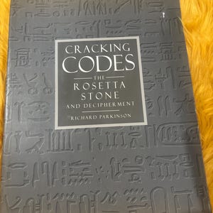 May include: A gray book titled Cracking Codes: The Rosetta Stone and Decipherment by Richard Parkinson. The cover features a dark gray background with embossed Egyptian hieroglyphs and a central rectangular panel containing white text.