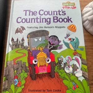 Könnte beinhalten: Ein Sesame Street Pop-up-Buch mit dem Titel The Count's Counting Book mit Jim Hensons Muppets. Das Cover zeigt den Grafen in einem lila-gelben Auto mit dem Nummernschild COUNT 1, ein Schloss, eine Fledermaus, einen Vogel und zwei Kaninchen.