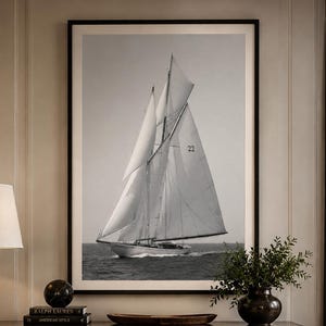 May include: A large, framed black and white wall art print featuring a sailboat with the number 22 on its sail, navigating across dark water. The artwork is displayed in a dark wooden frame and hangs above a dark wooden sideboard in a room.