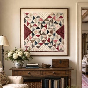 Könnte beinhalten: Ein quadratischer, an der Wand montierter Quilt mit einem geometrischen Muster aus Dreiecken in Rot-, Marineblau- und Cremetönen, gerahmt von einem einfachen Holzrahmen. Der Quilt hängt über einem hölzernen Konsolentisch mit Schubladen.