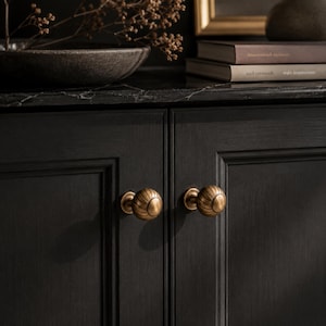 Peut inclure: Deux boutons de placard ronds en laiton avec une texture cannelée et nervurée verticale. Les boutons sont fixés sur une paire de portes de placard gris anthracite avec un grain de bois visible, dotées d'une construction à cadre et panneau.