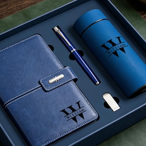 Könnte beinhalten: Ein blaues Geschenkset in einer Box mit einem Notizbuch mit Riemen, einem Kugelschreiber, einer zylindrischen Thermoskanne und einem USB-Stick. Das Notizbuch und die Thermoskanne tragen ein schwarzes Logo mit dem Namen William.