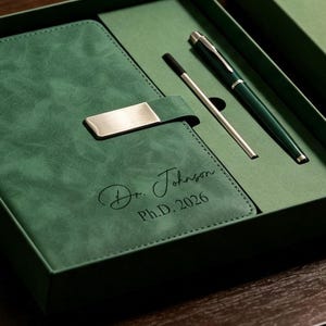 May include: A dark green notebook with a silver metal clasp and the engraved text Dr. Johnson Ph.D. 2026, accompanied by a matching dark green pen and a thin silver metal stylus, all presented within a dark green gift box.