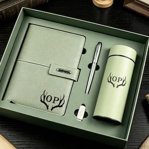 May include: A sage green gift set in a box containing a textured notebook with a strap closure, a silver pen, a small rectangular USB drive, and a matching sage green cylindrical insulated tumbler, all featuring a black antler and OP logo design.