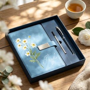 May include: A light blue notebook featuring a white daisy illustration and the name Kayla in script, secured with a silver metal clasp. The set includes a matching black ballpoint pen and a refill cartridge, all presented in a blue gift box.