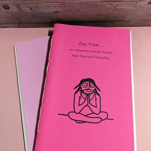 Puede incluir: Dos libros de tapa blanda. El libro superior es de color rosa brillante con el título "Zen Your..." y una ilustración en negro de una figura meditando. El libro inferior es de un tono rosa más claro. Los libros están sobre una superficie rosa.
