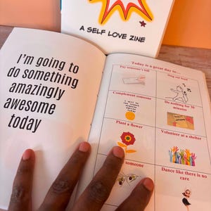 May include: An open book with the text "I'm going to do something amazingly awesome today" on the left page. The right page has illustrations and text, including "Today is a great day to..." and "A SELF LOVE ZINE" on the cover.