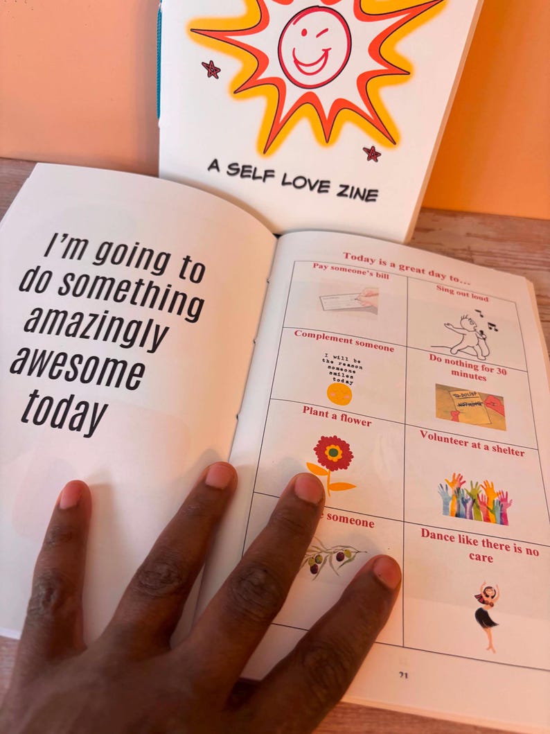 May include: An open book with the text "I'm going to do something amazingly awesome today" on the left page. The right page has illustrations and text, including "Today is a great day to..." and "A SELF LOVE ZINE" on the cover.