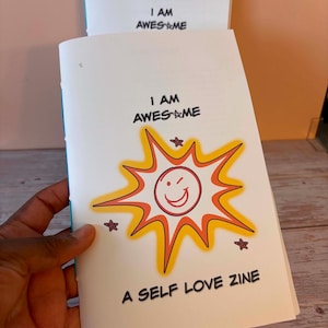 Op de afbeelding: Een stapel zelfliefde zines met de tekst "I AM AWESOME" en "A SELF LOVE ZINE". De omslag heeft een lachend zonnestraalontwerp in oranje, geel en rood met steraccenten. De zines worden in een hand gehouden.