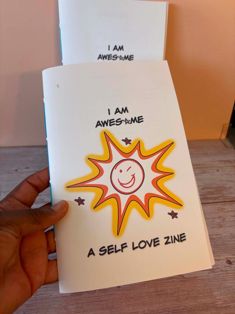 May include: A stack of self-love zines with the text "I AM AWESOME" and "A SELF LOVE ZINE". The cover features a smiling sunburst design in orange, yellow, and red with star accents. The zines are held in a hand.