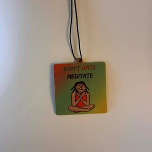 Może przedstawiać: Kwadratowa drewniana ozdoba z czarnym sznurkiem. Ozdoba ma gradientowe tło w kolorach czerwonym, ż&oacute;łtym i zielonym. Tekst "DON'T HATE MEDITATE" jest wydrukowany nad postacią z kresk&oacute;wki w pozycji jogi.