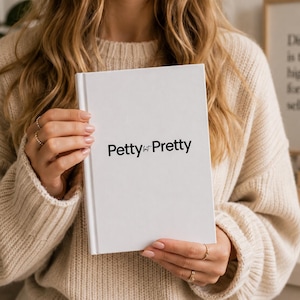 May include: A white book with the text "Petty for Pretty" in black lettering. The book is held by a person wearing a beige sweater. The background includes a framed quote and a lit candle.